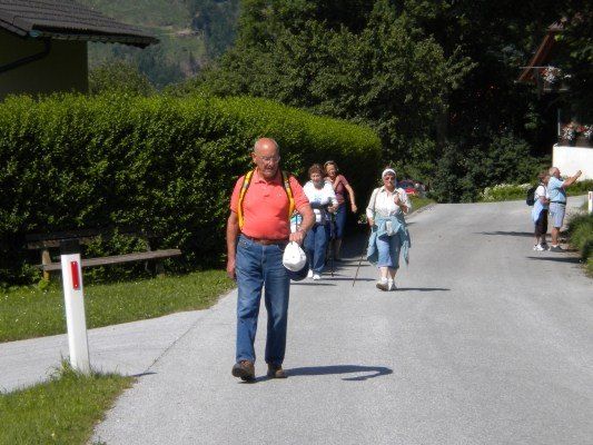 Eine Gruppe älterer Menschen geht auf einem Pfad in einem Dorf. Der Mann vorne trägt ein rotes Hemd und trägt eine weiße Tasche.