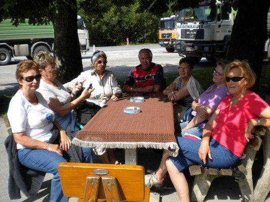 Eine Gruppe älterer Frauen und Männer sitzt um einen Tisch in einem Park, mit geparkten Lastwagen im Hintergrund.
