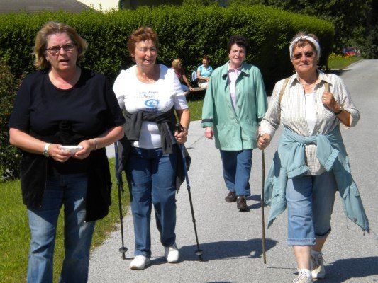 Vier Frauen gehen auf einem gepflasterten Weg, zwei von ihnen benutzen Gehstöcke. Sie sind in Freizeitkleidung gekleidet, und eine trägt eine weiße Kopfbinde. Hinter ihnen befinden sich Büsche und ein Rasenbereich, auf dem einige Leute sitzen.