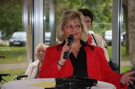Eine Frau in einem roten Mantel spricht in ein Mikrofon bei einer Veranstaltung im Freien, mit einer weiteren Frau im Hintergrund und einem Tisch mit Papieren im Vordergrund.
