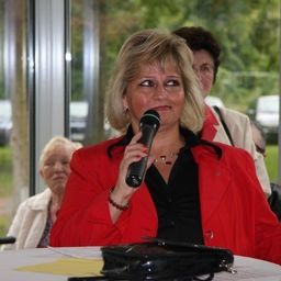 Eine Frau in einem roten Mantel spricht in ein Mikrofon bei einer Veranstaltung im Freien, mit einer weiteren Frau im Hintergrund und einem Tisch mit Papieren im Vordergrund.