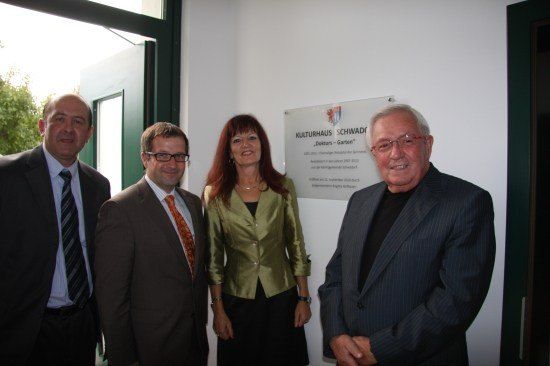 Vier Personen in formeller Kleidung stehen zusammen vor einer weißen Wand mit einem Schild, das 'Kulturhaus SCHWADI' und 'Deiters-Garten' besagt. Der Mann auf der linken Seite trägt eine gestreifte Krawatte, während die Frau in der Mitte eine Halskette hat. Der Mann auf der rechten Seite trägt eine Brille.