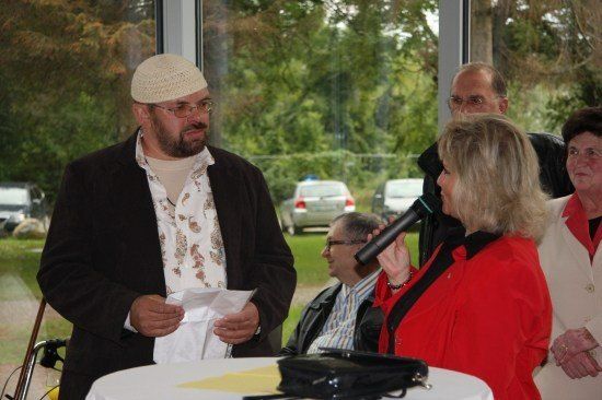 Ein Mann mit einer weißen Mütze liest von einem Stück Papier, während eine Frau in einem roten Mantel ein Mikrofon hält. Hinter ihnen sitzen zwei weitere Personen und beobachten. Draußen am Fenster sind Autos und Bäume zu sehen.