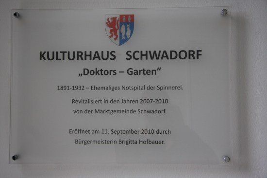 Ein Schild an einer Wand zeigt 'Kulturhaus Schwadorf' mit einem Löwen- und Fischemblem darüber. Es erwähnt, dass das Gebäude von 1891 bis 1932 ein ehemaliges Krankenhaus war. Es wurde in den Jahren 2007-2010 von der Schwadorf-Gemeinde wiederbelebt und am 11. September 2010 eröffnet.