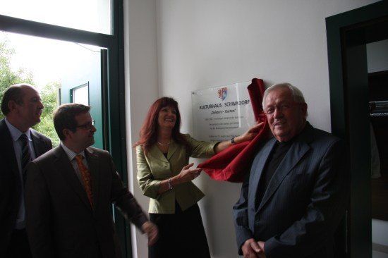 Eine Gruppe von Menschen, darunter eine Frau, die ein rotes Band hält, steht vor einer weißen Wand mit einem Schild. Der Mann rechts lächelt.