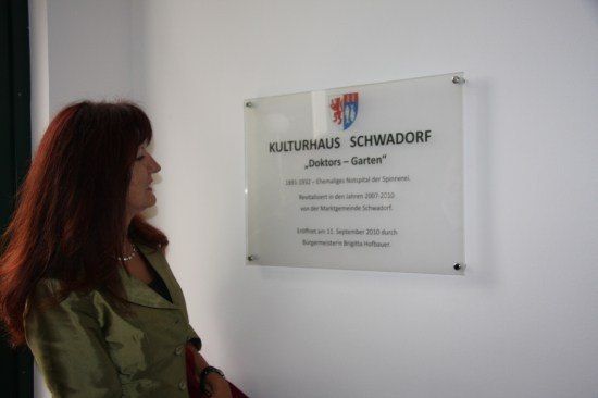 Eine Frau mit roten Haaren steht vor einem Schild an einer weißen Wand. Das Schild heißt 'Kulturhaus Schwadorf' und enthält ein Logo und Text.