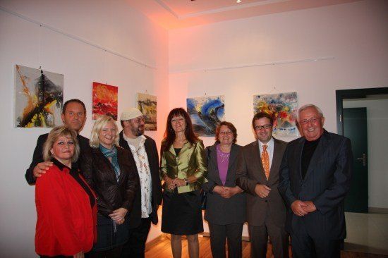 Eine Gruppe von Menschen steht in einer Kunstgalerie, lächelt und posiert für ein Foto. Der Raum hat weiße Wände und Holzböden. An den Wänden hängen mehrere Gemälde.