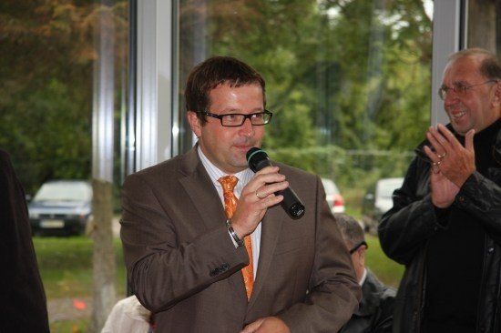 Ein Mann in einem braunen Anzug und einer orangefarbenen Krawatte spricht in ein Mikrofon, während ein anderer Mann hinter ihm klatscht.