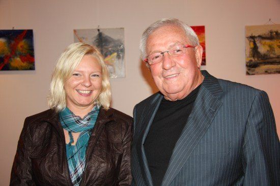Ein älterer Mann und eine Frau stehen zusammen vor einer Wand mit abstrakten Gemälden. Die Frau lächelt, während der Mann eine Brille und einen Anzug trägt.