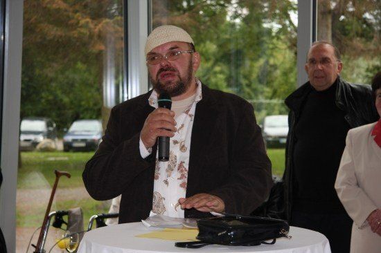 Ein Mann mit Bart und weißer Kappe spricht in ein Mikrofon bei einer Veranstaltung, bei der zwei weitere Personen hinter ihm stehen.