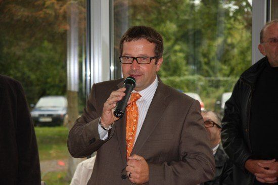 Ein Mann in einem braunen Anzug und einer orangefarbenen Krawatte spricht in ein Mikrofon bei einer Veranstaltung im Freien. Hinter ihm steht ein Mann in einem schwarzen Jackett, der aufmerksam zuhört.