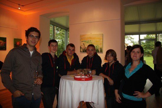 Eine Gruppe von Menschen, darunter Männer und Frauen, steht um einen Tisch mit Weinläsern und einer roten Schale, möglicherweise bei einer gesellschaftlichen Veranstaltung.