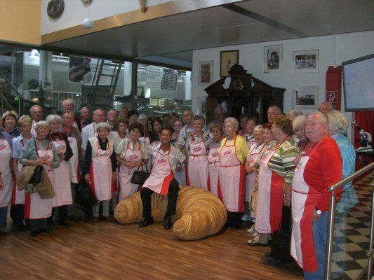 Eine Gruppe älterer Menschen in Schürzen versammelt sich in einem Raum, einige stehen hinter einer großen Croissant-Skulptur. Sie scheinen für ein Foto zu posieren.