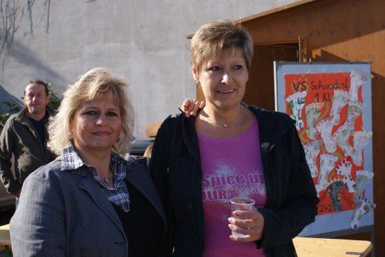 Zwei Frauen stehen eng beieinander und lächeln für ein Foto. Eine hält einen Becher, die andere hat ihren Arm um die erste gelegt. Hinter ihnen steht ein Mann in der Nähe einer Wand mit einem Plakat.