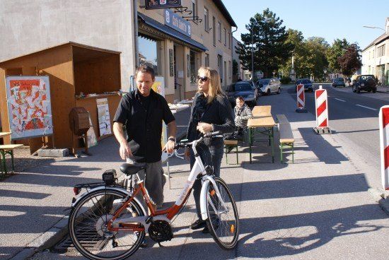 Zwei Personen stehen neben einem Fahrrad auf der Straße, mit einem Café und Sitzgelegenheiten im Hintergrund.