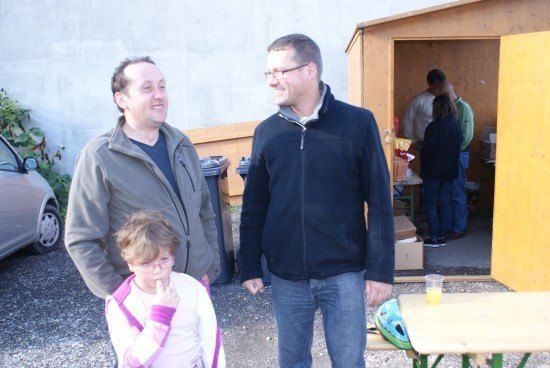 Drei Personen stehen vor einem kleinen Schuppen. Zwei Männer lächeln und ein Mädchen hält sich den Finger an den Mund.