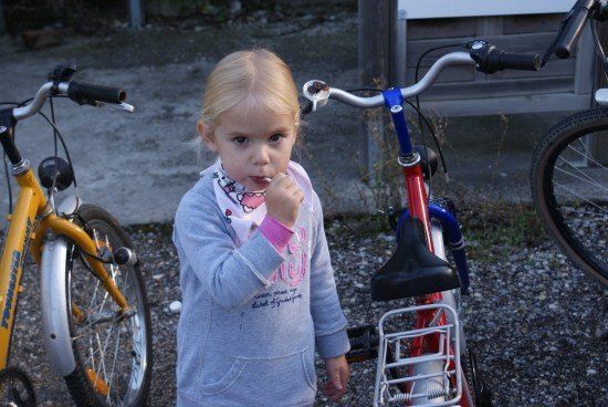 Ein kleines Mädchen mit blonden Haaren steht vor einem roten Fahrrad und hält eine Zahnbürste in der Hand. Sie trägt ein graues Sweatshirt mit einem rosa Kragen. Hinter ihr stehen zwei Fahrräder und ein Gebäude.