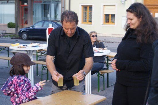 Ein Mann gießt Getränke in zwei Gläser, während eine Frau und ein junges Mädchen zusehen. Sie stehen vor einem Tisch mit Bänken und einem geparkten Auto in der Nähe.