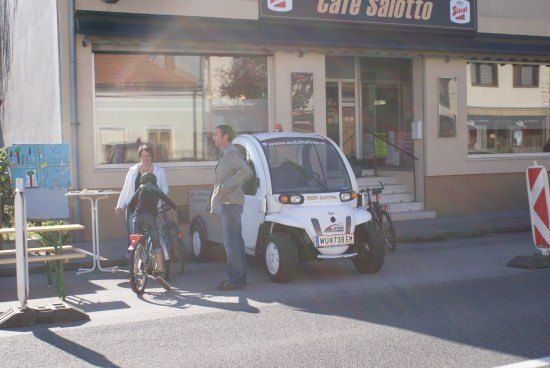 Zwei Personen stehen neben einem kleinen Elektroauto vor einem Café. Der Mann spricht mit der Frau, die ein Fahrrad hält.