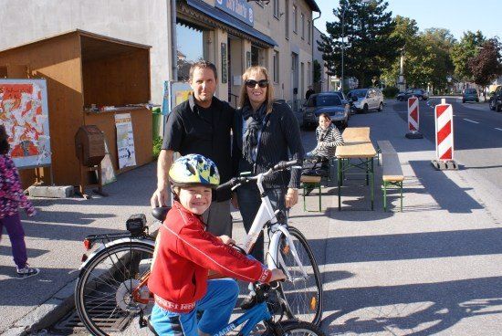 Ein Mann und eine Frau stehen neben einem Jungen auf einem Fahrrad, mit einem Geschäft und geparkten Autos im Hintergrund.