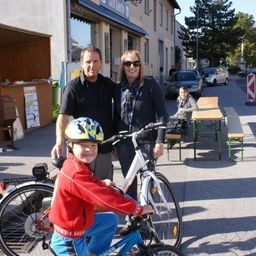 Ein Mann und eine Frau stehen neben einem Jungen auf einem Fahrrad, mit einem Geschäft und geparkten Autos im Hintergrund.
