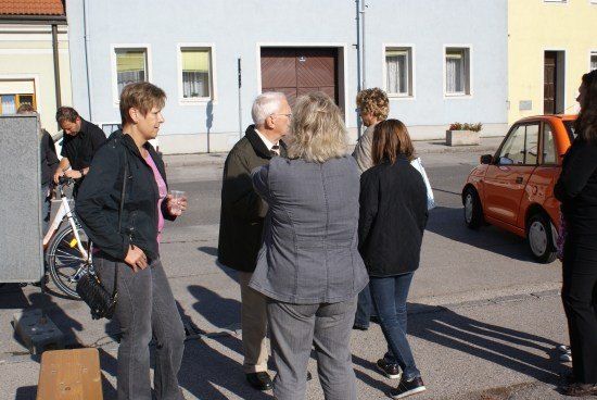 Eine Gruppe von Menschen steht auf einer Straße, mit Gebäuden und einem Fahrrad im Hintergrund. Ein orangefarbenes Auto ist in der Nähe geparkt.