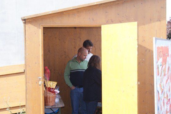 Drei Menschen stehen in einem Holzschuppen mit einer gelben Tür. Der Mann in der Mitte hat die Hände in den Taschen.