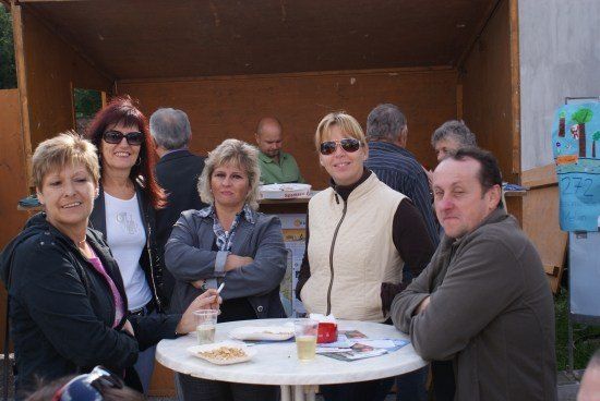Eine Gruppe Erwachsener, einige mit Sonnenbrillen, versammelt sich um einen Tisch mit Essen und Getränken, möglicherweise bei einer Outdoor-Veranstaltung.