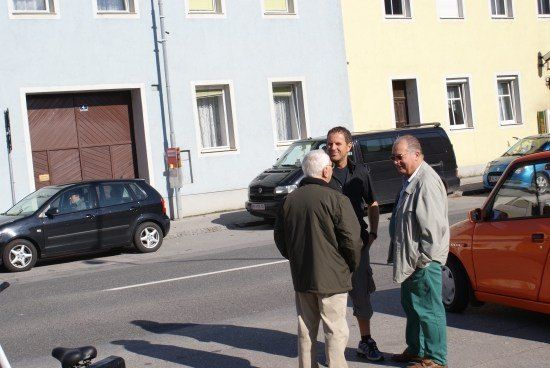 Drei Männer stehen an einer Straßenecke, mit einem schwarzen Auto hinter ihnen und einem orangefarbenen Auto auf der rechten Straßenseite.