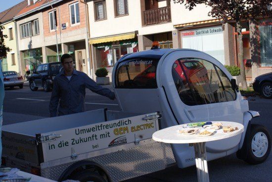 Ein Mann steht neben einem kleinen Elektrofahrzeug mit einem Tisch voller Essen davor. Das Fahrzeug hat ein Schild mit der Aufschrift Umweltfreundlich in die Zukunft mit GEM ELECTRIC.