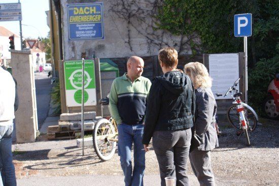 Drei Personen stehen auf einer Straße, ein Mann in der Mitte trägt ein grünes Shirt und blaue Jeans. Ein Fahrrad ist am Straßenrand geparkt. Hinter ihnen befindet sich ein Gebäude mit einem Schild, das Dach Ehrenberger liest.