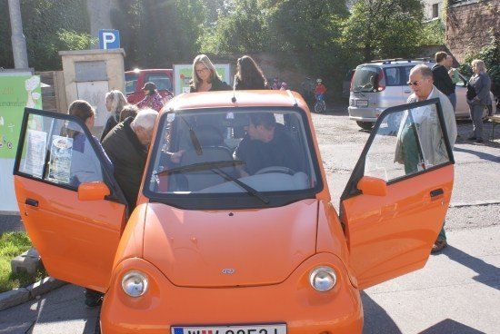Ein orangefarbenes Auto mit geöffneten Türen ist am Straßenrand geparkt. Mehrere Menschen sind um es herum versammelt, darunter ein Mann, der im Inneren sitzt.