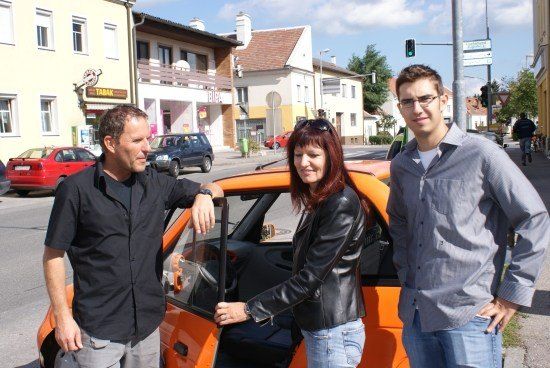 Drei Personen stehen neben einem orangefarbenen Auto auf einer Straße mit Gebäuden und geparkten Autos im Hintergrund.