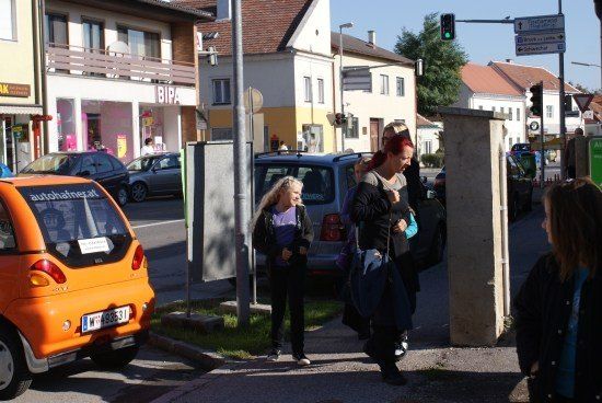 Mehrere Personen gehen auf einem Bürgersteig in der Nähe einer belebten Straße mit geparkten Autos und Gebäuden im Hintergrund. Eine Frau trägt eine Tasche.