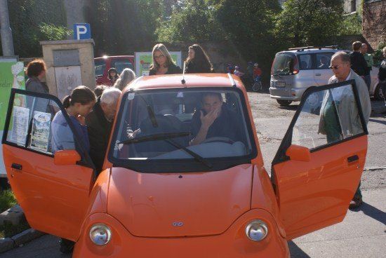 Ein Mann sitzt in einem orangefarbenen Auto mit offenen Türen, während mehrere Personen um ihn herumstehen. Im Hintergrund befindet sich ein Parkzeichen und ein roter Pickup-Truck.
