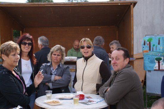 Eine Gruppe von Menschen versammelt sich um einen Tisch, möglicherweise bei einer sozialen Veranstaltung. Eine Frau raucht eine Zigarette und eine andere Frau lächelt. Auf dem Tisch befinden sich Teller, Gläser und andere Gegenstände. Im Hintergrund steht eine Holzkonstruktion mit einem Plakat.