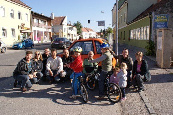 Eine Gruppe von Menschen, einschließlich Kindern, posiert für ein Foto auf einer Straße mit Gebäuden und geparkten Autos im Hintergrund.