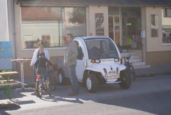 Ein Mann und eine Frau stehen neben einem kleinen weißen Elektroauto, vor dem ein Kind auf einem Fahrrad fährt, vor einem Gebäude.