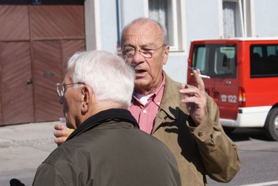 Zwei ältere Männer stehen draußen, einer raucht eine Zigarette und der andere hält ein Getränk. Sie stehen vor einem roten Van und einem Gebäude.