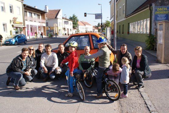 Eine Gruppe von Menschen, einschließlich Kindern, posiert für ein Foto am Straßenrand. Einige tragen Helme und halten Fahrräder. Im Hintergrund befindet sich ein Gebäude mit einem Schild.