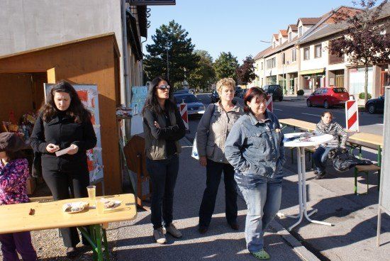 Fünf Frauen stehen auf einem Bürgersteig, zwei mit verschränkten Armen, in der Nähe eines Tisches mit Essen. Eine weitere Frau sitzt hinter ihnen an einem Tisch. Gebäude und geparkte Autos säumen die Straße.