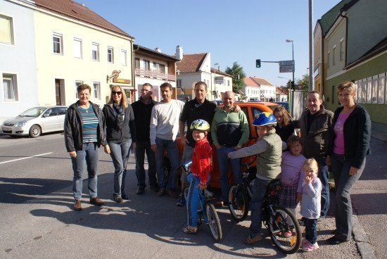 Eine Gruppe von Menschen, einschließlich Kinder mit Fahrrädern, posiert für ein Foto auf einer Straße mit Gebäuden und geparkten Autos im Hintergrund.