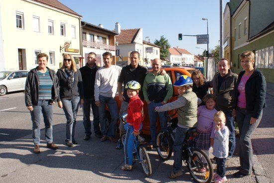 Eine Gruppe von Erwachsenen und Kindern, einige mit Helmen, stehen auf einer Straße mit Fahrrädern und posieren für ein Foto vor Gebäuden und einer Ampel.