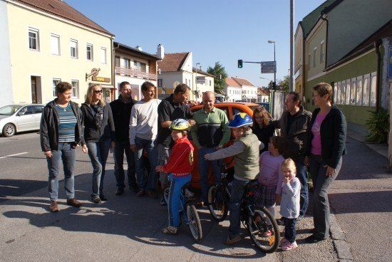 Eine Gruppe von Erwachsenen und Kindern steht an einer Straßenecke, einige mit Fahrrädern, unter einem klaren Himmel. Gebäude säumen die Straße, und eine Ampel ist sichtbar.