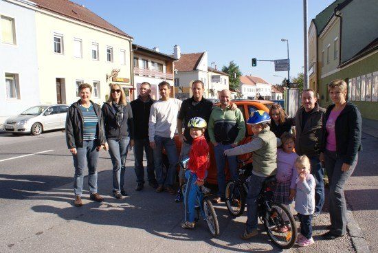 Eine Gruppe von Menschen, darunter Erwachsene und Kinder, steht mit Fahrrädern auf der Straße. Sie alle lächeln und posieren für ein Foto. Hinter ihnen befinden sich Gebäude, ein geparktes Auto und eine Ampel.