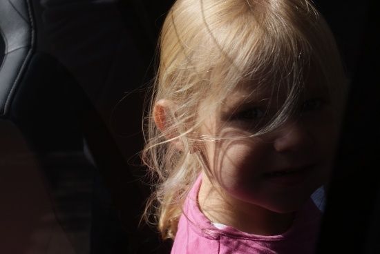 Ein kleines Mädchen mit blonden Haaren lächelt und trägt ein rosa T-Shirt. Sie steht in einem schwach beleuchteten Bereich.