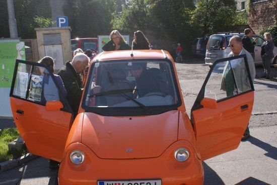 Ein kleines orangefarbenes Auto mit geöffneten Türen, das am Straßenrand geparkt ist, mit Menschen, die herumstehen.