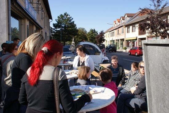Eine Gruppe von Menschen sitzt um einen Tisch herum vor einem Gebäude, einige essen, andere unterhalten sich. Am Straßenrand sind Autos geparkt.