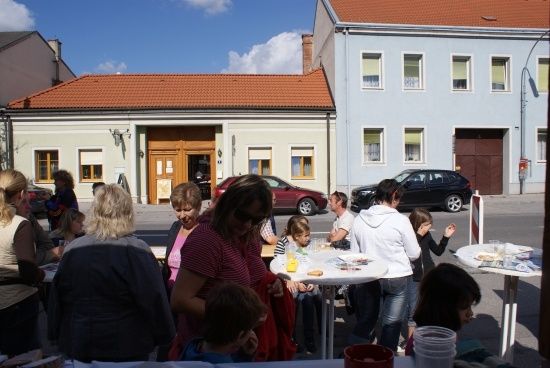 Eine Gruppe von Menschen, einschließlich Kindern, versammelt sich an Tischen vor einem Gebäude an einem sonnigen Tag.
