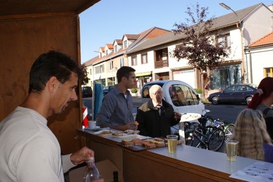 Ein Mann in einem weißen Shirt steht an einem Tresen und hält eine Flasche. Hinter ihm spricht ein Mann in einem blauen Shirt mit einem älteren Mann in einem schwarzen Mantel. In der Nähe sind Fahrräder geparkt.